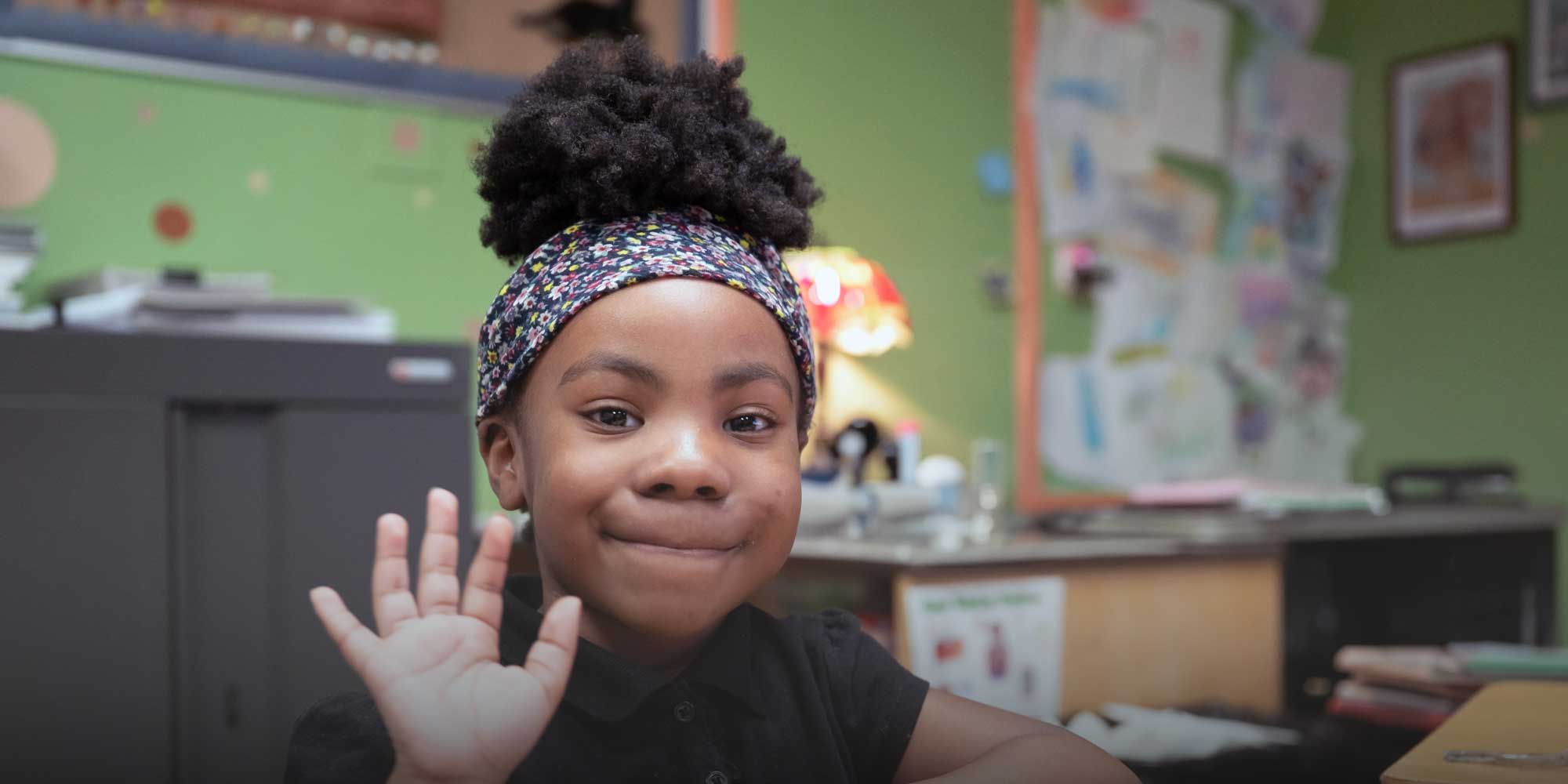 Young student in a floral headband waving in a classroom.