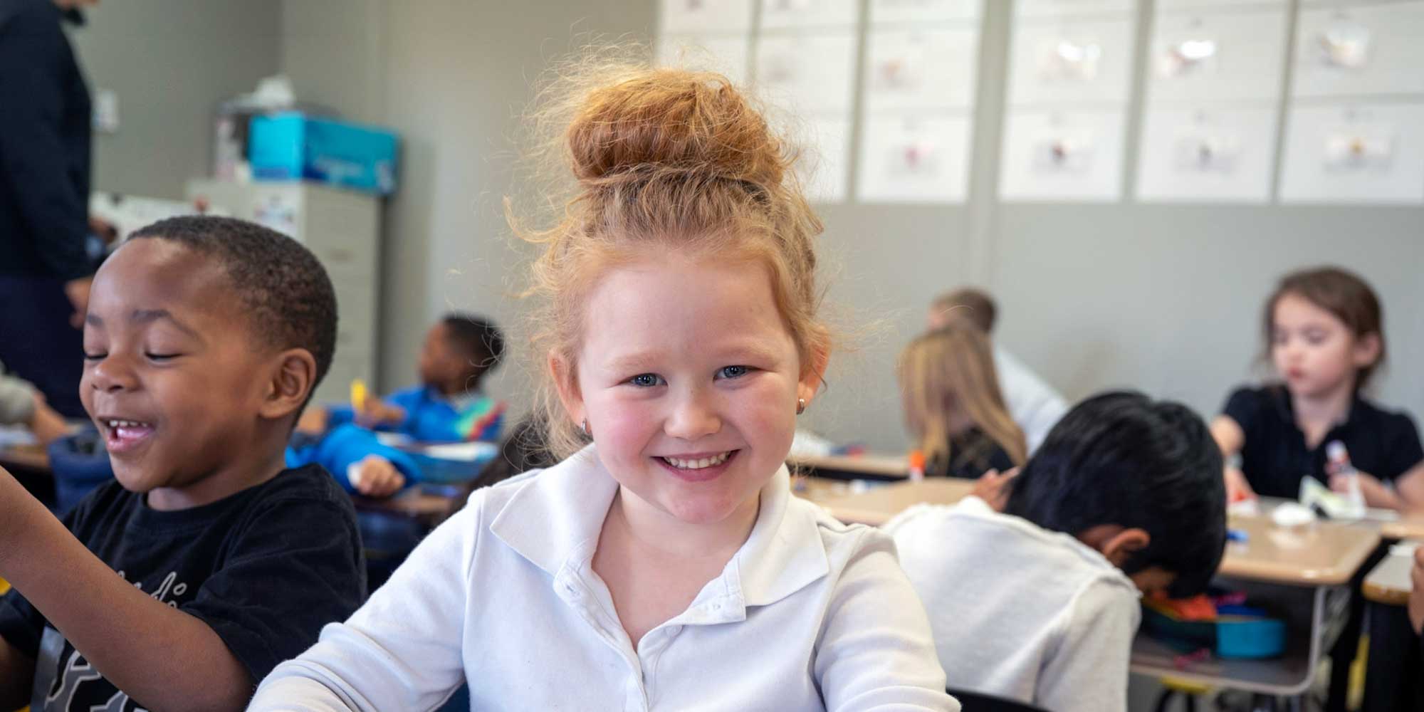 Smiling student at her desk in a busy classroom.